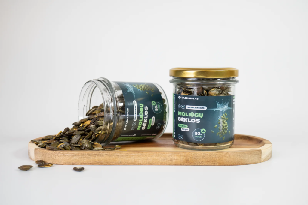 Two glass jars of pumpkin seeds on a wooden tray, one tipped over with seeds spilled onto the surface and to the side.