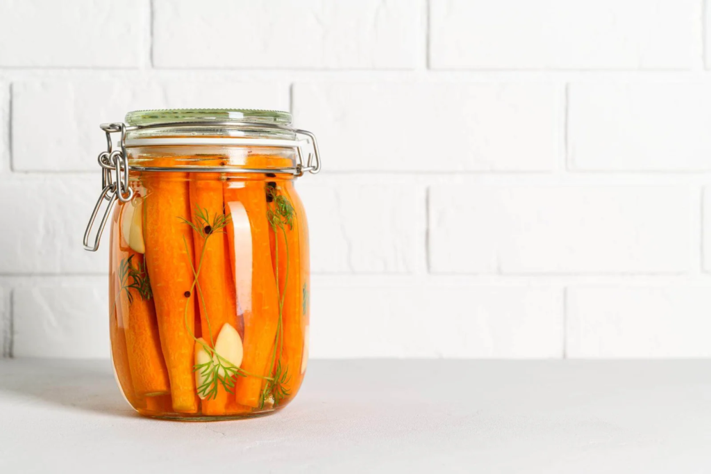 Jar of pickled carrot spears with garlic cloves and dill in brine on a white surface and brick backdrop